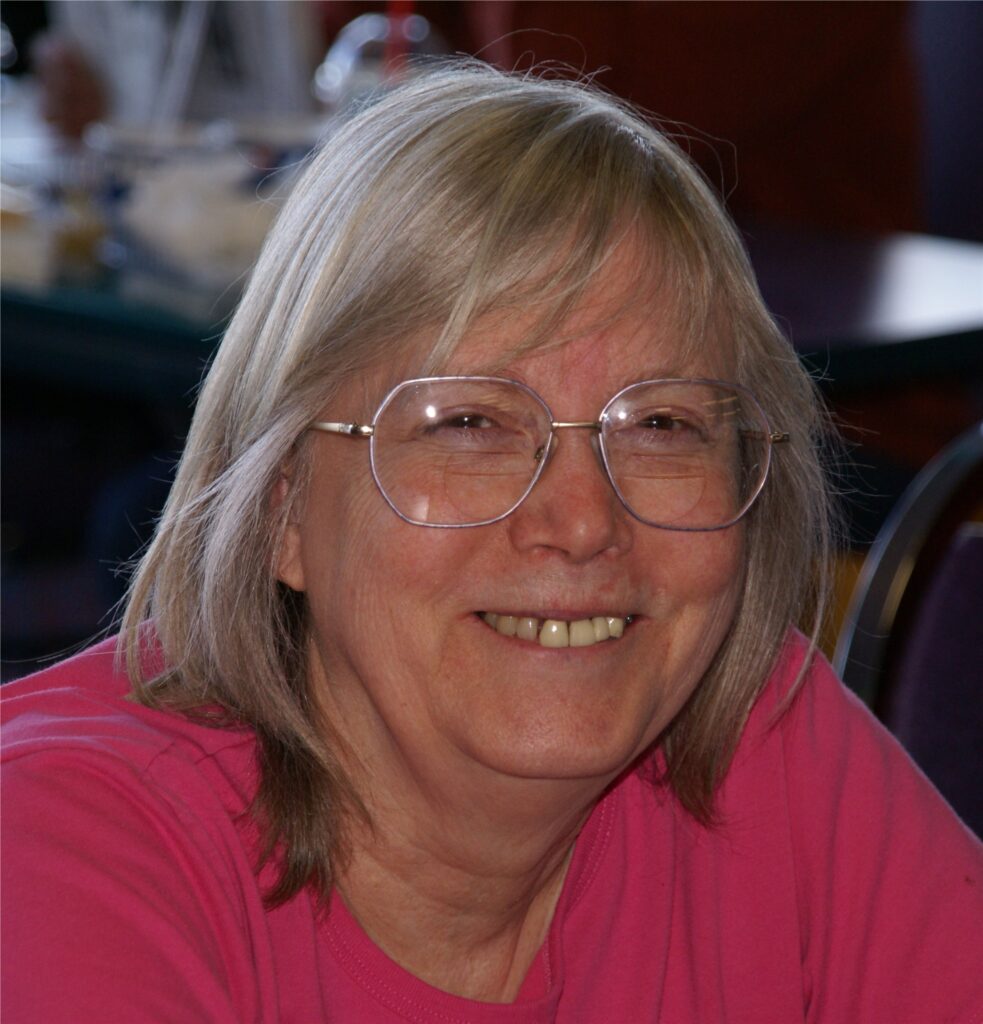 A white woman smiles with sandy grey hair that goes to her shoulders, wearing large hexagon shaped glasses and a vivid hot pink shirt. She looks very happy. The background is blurred and appears to be a restaurant or some sort of gathering.